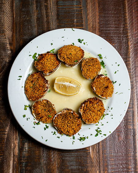 Clams Oreganato presented as 8 littleneck clams baked with italian seasoned breadcrumbs plated on a white round dish and placed on a wooden table