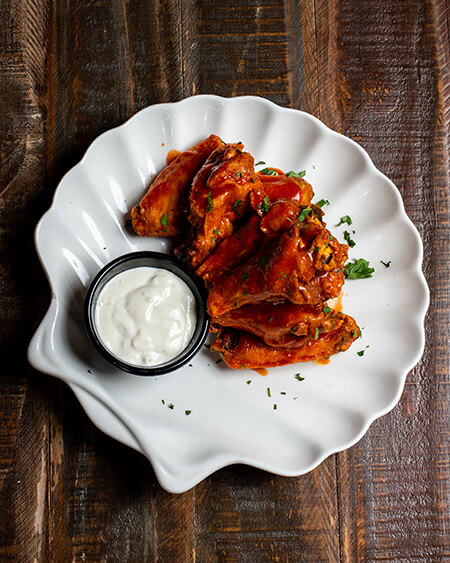 Buffalo Wings presented as 8 piece jumbo party wings with blue cheese plated on a white shell plate and placed on a wooden table