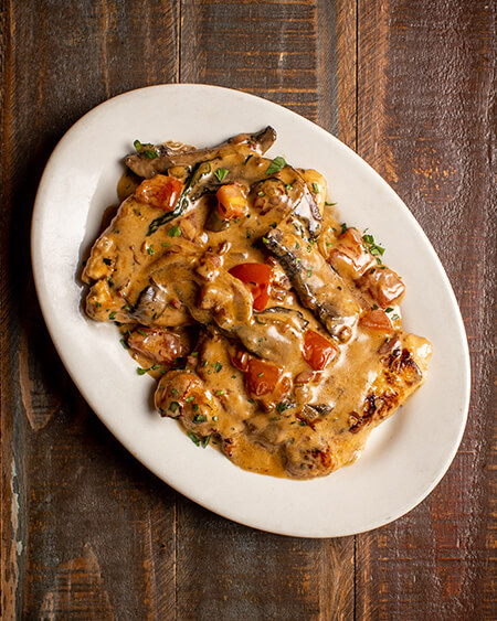 Chicken Boscaiola presented as 2 sautéed chicken breasts with portobello mushrooms, diced tomatoes, shallots, and prosciutto in brandy cream sauce with a touch of demi-glaze plated on a round white dish and placed on a wooden table