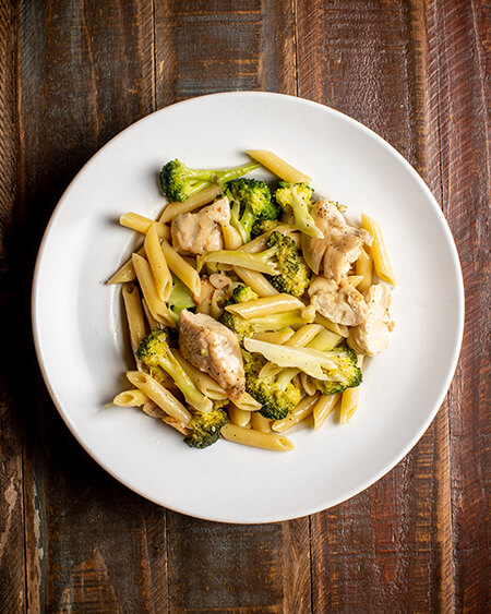 A beautifully presented plate of Cavetelli, Chicken, and Broccoli pasta on a wooden counter — $15.95