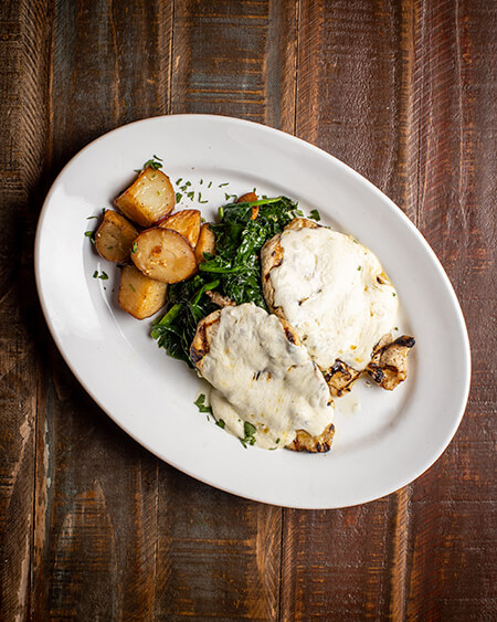 Chicken Florentine presented as 2 grilled chicken breasts topped with fresh mozzarella cheese over spinach and roasted potatoes plated on a white dish and placed on a wooden table