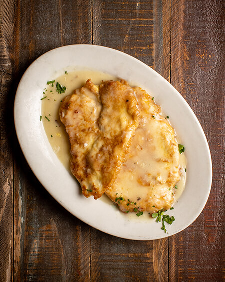 Francese Chicken presented as 2 pieces of egg battered chicken breast sautéed in francese sauce (lemon-butter and white wine sauce) plated on a white round dish and placed on a wooden table