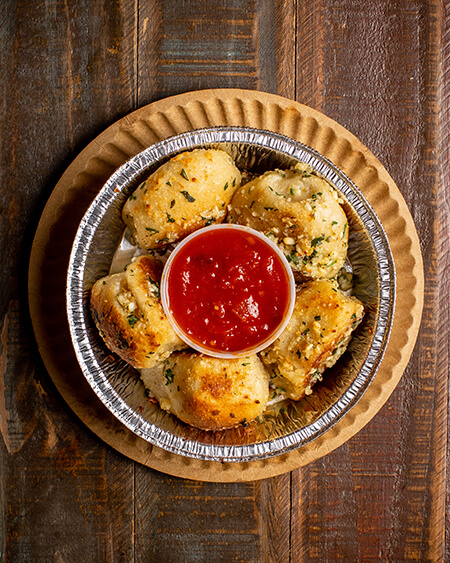 Garlic Knots presented as 5 garlic knots with a side of marinara sauce plated in a tin dish on a wooden table