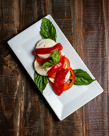 Mozzarella caprese salad is 3 slices of fresh mozzarella cheese, 3 slices tomato, roasted peppers and basil, extra virgin olive oil