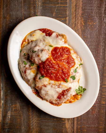 A beautifully presented plate of Parmigiana Chicken on a wooden counter — $12.50