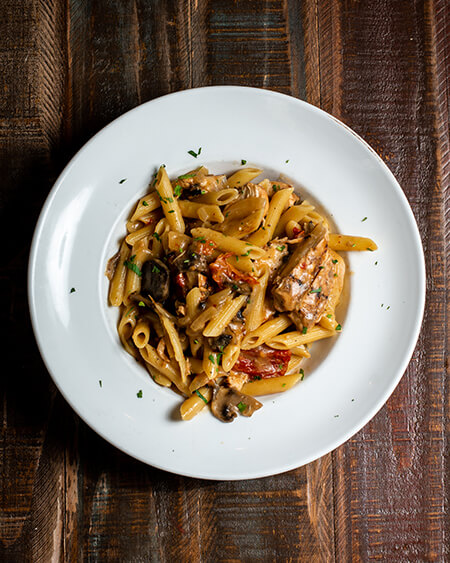 A beautifully presented plate of Penne con Pollo on a wooden counter — $13.95