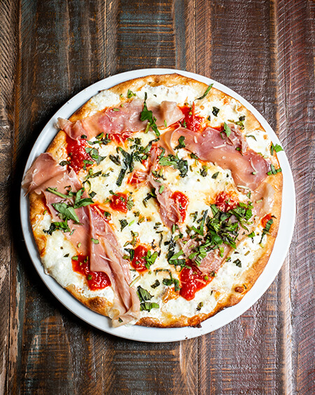 A beautifully presented personal gourmet prosciutto pizza on a wooden counter — $10.95