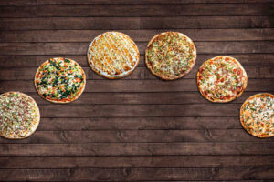 An array of six pizza pies from Renato's Pizza Masters sitting on a wooden countertop.