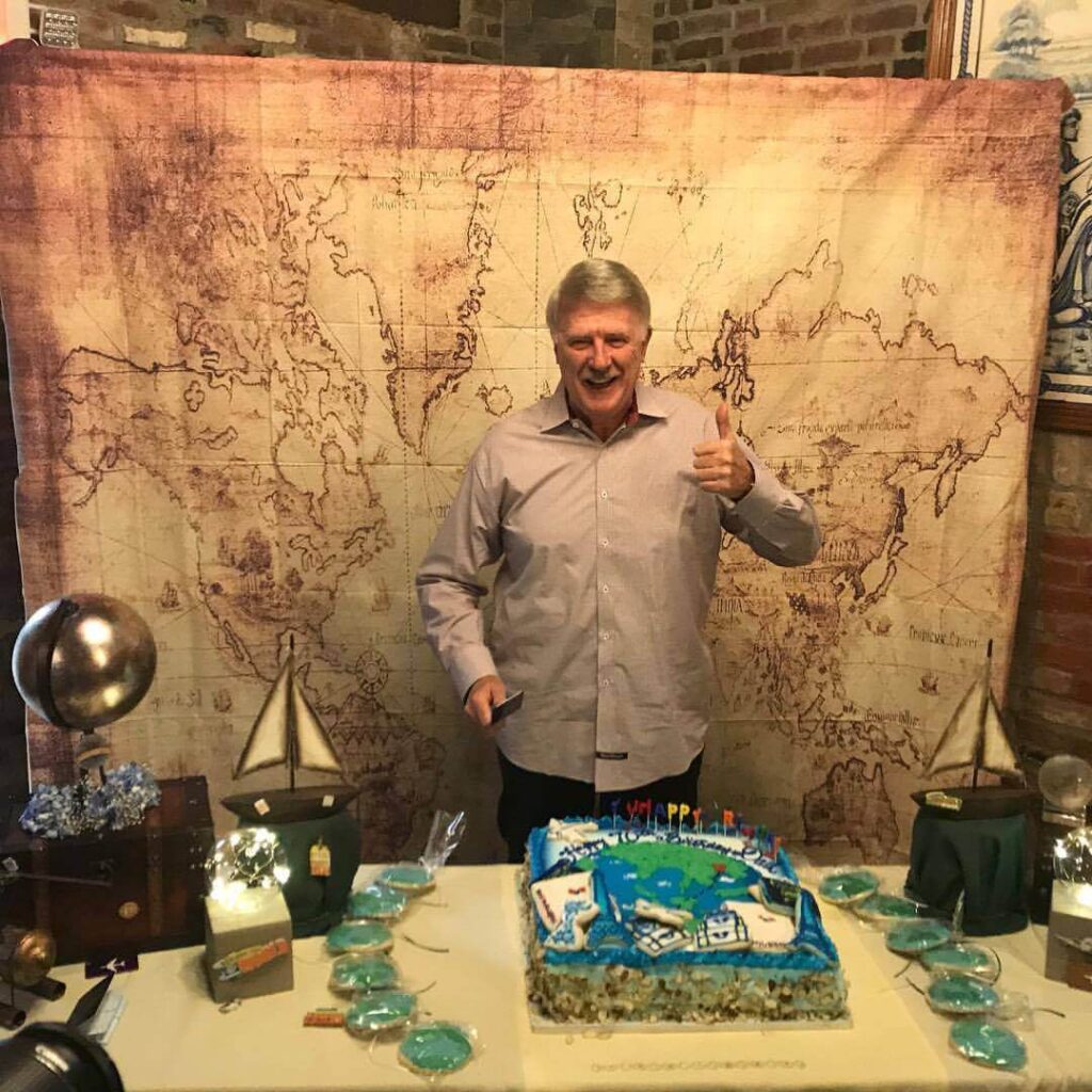 Richie DeMarchi standing in front a vintage map and behind of a celebration array including an anniversary cake.