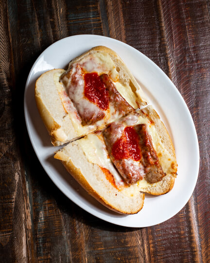 A beautifully presented Sausage Parmigiana Hot Sandwich on a wooden counter — $9.50