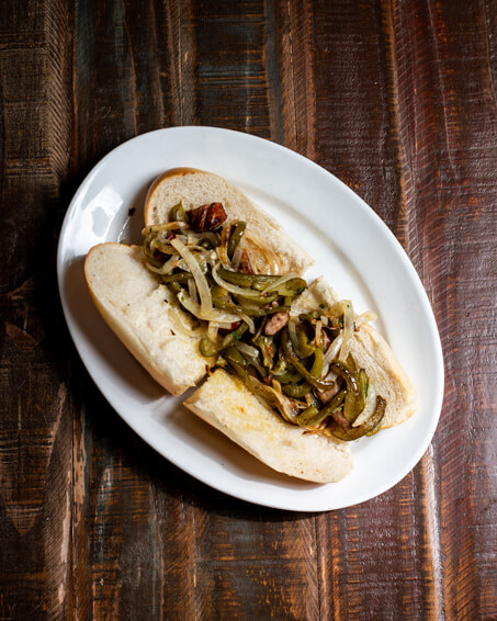 A beautifully presented Sausage, Peppers, and Onions Hot Sandwich on a wooden counter — $9.50