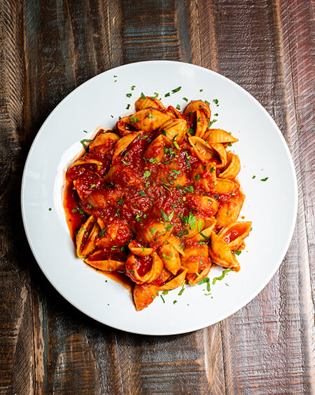Tomato Ragu Sauce Pasta presented as shells pasta in our signature tomato ragu sauce plated in a white round dish and placed on a wooden table