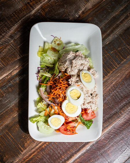 Tuna Salad is a Small house salad topped with tuna salad and a hard-boiled egg with house dressing displayed on a long rounded white dish on a wooden table